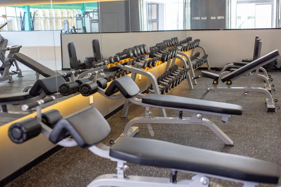 A quiet gym floor with rows of dumbbells and benches in front of a mirror