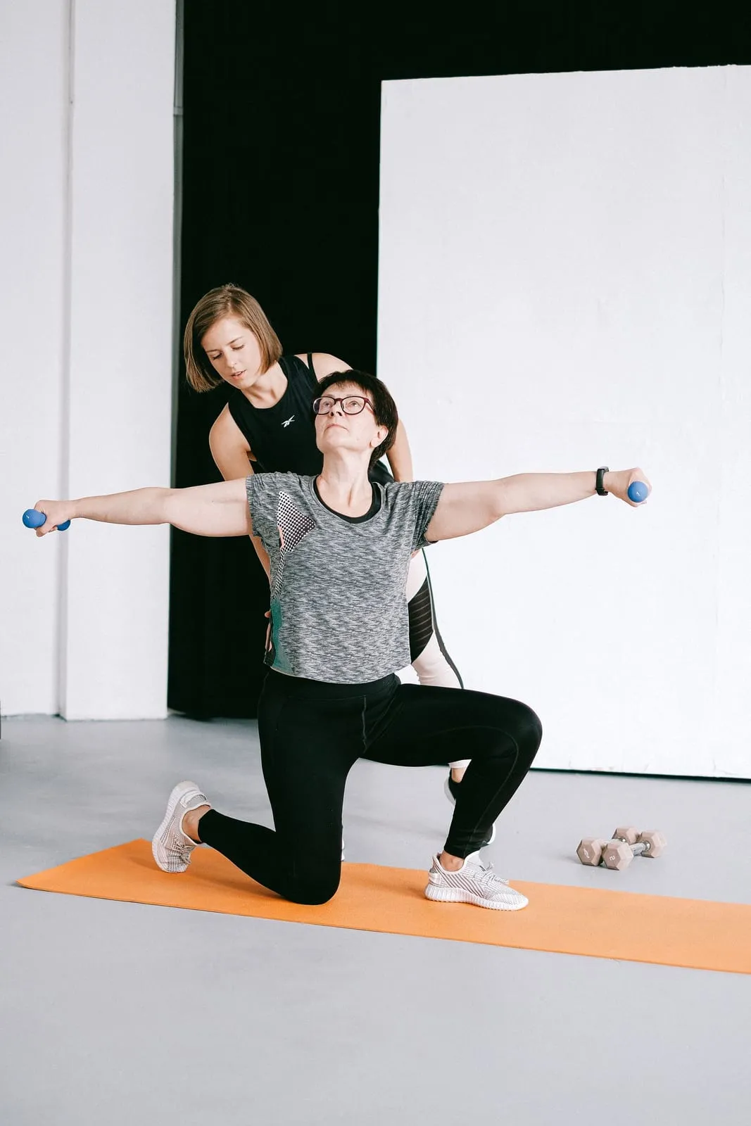 Helen in a kneeling lunge raising dumbbells to the side, her trainer behind her