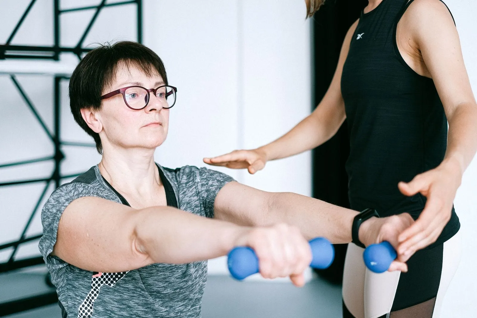 Helen raising light dumbbells in front of her while her trainer watches