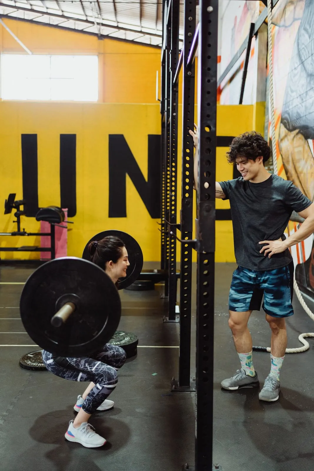 Sagar standing by a loaded barbell in the rack, listening to a trainer