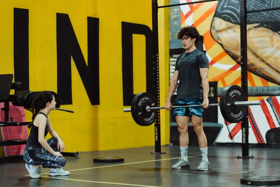 A female trainer crouching to watch a man finish a barbell lift in a gym